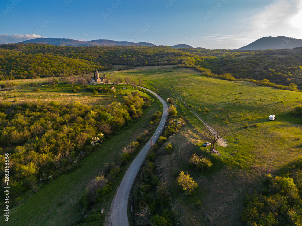 Fototapeta premium Ancient church in a mountains area and a road between green hills, aerial view