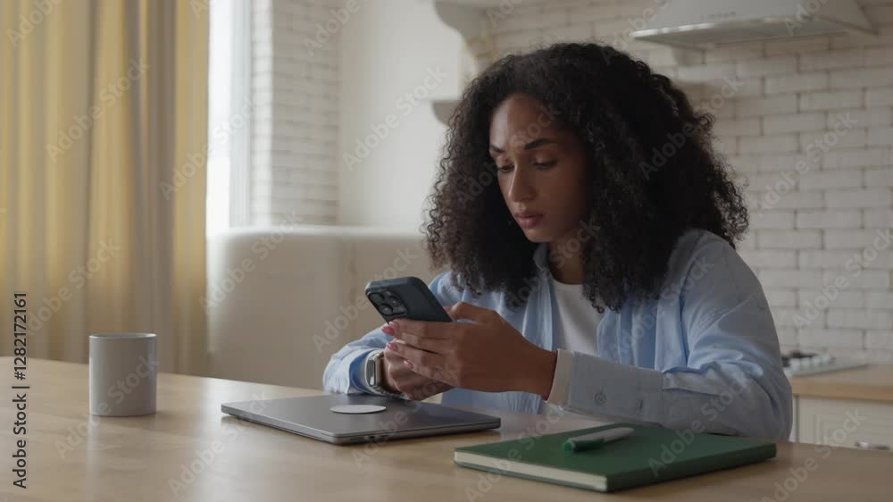 Young African Woman Sitting In Kitchen, Looking At Phone, Upset By Bad News Young African Woman Sitting In Kitchen, Looking At Phone, Upset By Bad News