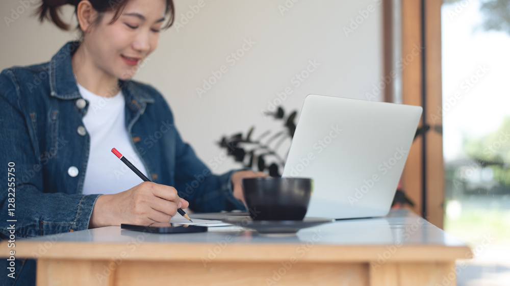 Asian woman working on laptop computer and writing on notepad on table at coffee shop. Smiling freelancer online working, planning new project, freelance at work Asian woman working on laptop computer and writing on notepad on table at coffee shop. Smiling freelancer online working, planning new project, freelance at work