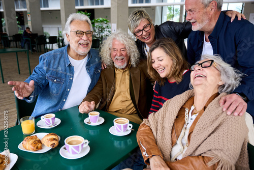 Cheerful group of mature people laughing and enjoying breakfast sitting at cafeteria bar table together. Retired generation community friends having fun gathered on terrace for snacking
