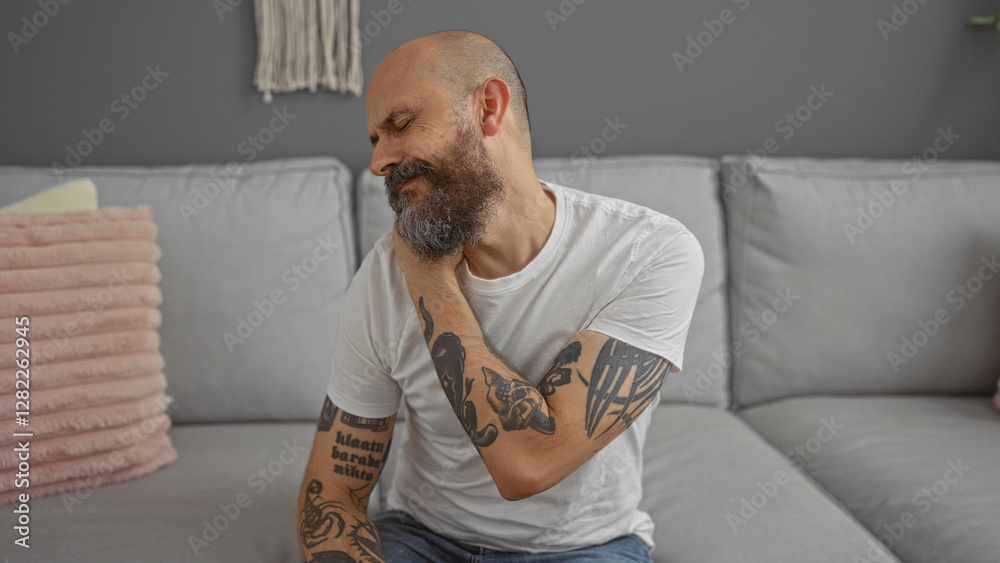 Handsome man with a beard and tattoos feeling neck pain in a modern living room setting with stylish decor, expressing discomfort while sitting on a comfortable couch at home Handsome man with a beard and tattoos feeling neck pain in a modern living room setting with stylish decor, expressing discomfort while sitting on a comfortable couch at home