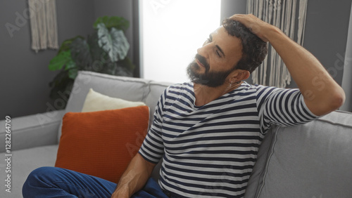 Canvas Print Young hispanic man stretches neck in a cozy living room, seated on a comfortable sofa with decorative pillows and a relaxed, thoughtful demeanor indoors