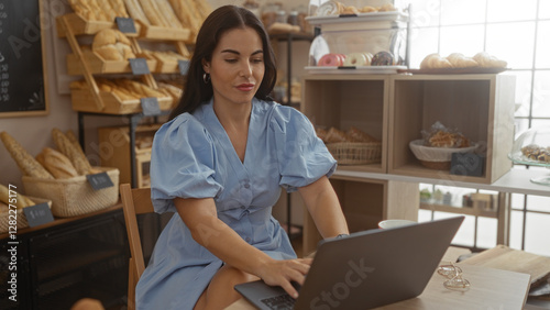 Young woman working inside a bakery on a laptop, surrounded by bread and pastries, appears focused and engaged in a cozy, well-lit cafe setting