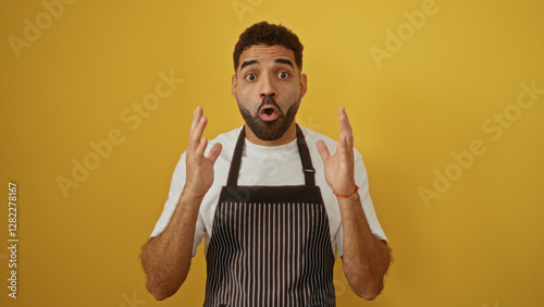 Young hispanic man in an apron expressing surprise while standing against an isolated yellow background.