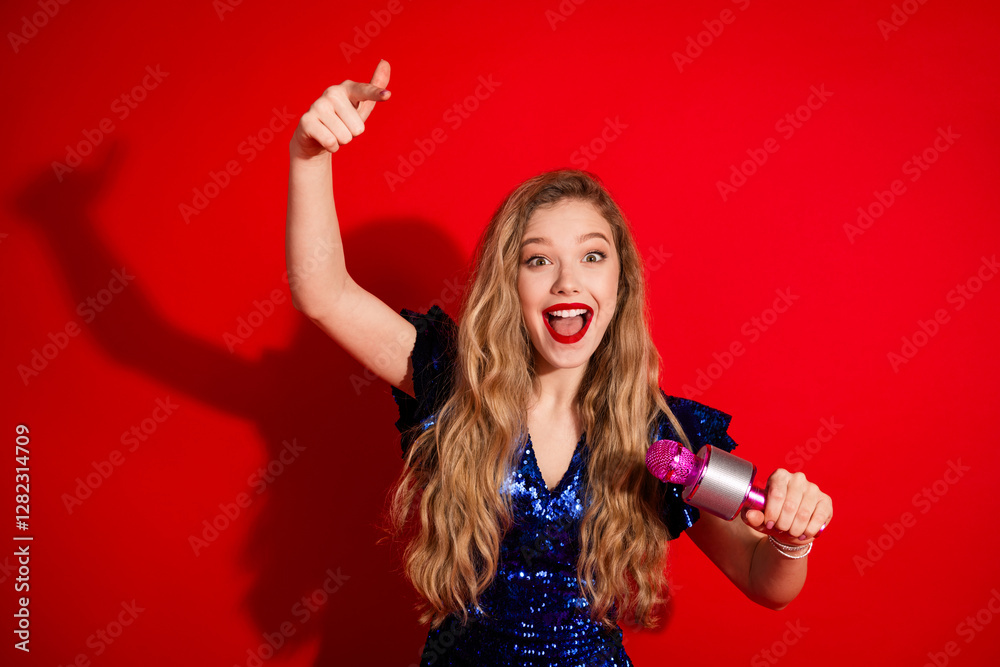 Fototapeta premium Cheerful young woman in a shiny blue dress posing with a microphone on a vibrant red background