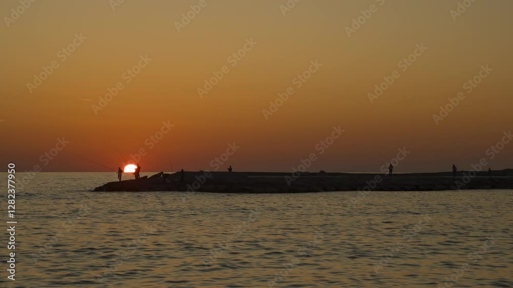 Zoom in Time Lapse of Fishermen with Fishing Rods Standing on a Rocky Pier During Sunset over the Sea with an Orange Sky in Silhouette Zoom in Time Lapse of Fishermen with Fishing Rods Standing on a Rocky Pier During Sunset over the Sea with an Orange Sky in Silhouette