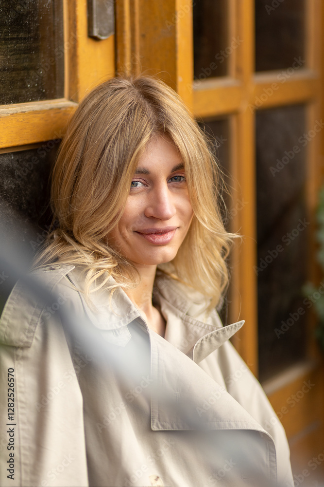 Fototapeta premium Woman with blonde hair relaxing by a yellow door while wearing a light trench coat in a cozy urban setting