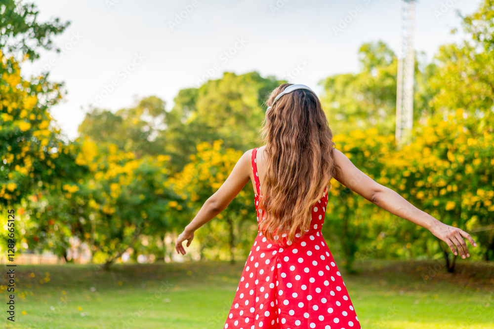 Obraz premium Young Woman in Retro Red Polka Dot Dress Wearing Headphones Listening and Enjoying Music in a Park Happily, Immersing with Nature