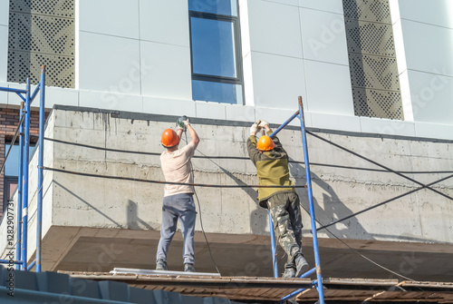 Workers standing on scaffolding work with hand-held power tools. Facade work at a height. Builders are drilling the facade of the building for the cladding of the building. Construction of buildings.