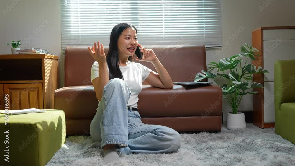 Relaxed young woman sitting on floor, engaging in phone conversation while making expressive hand gestures within modern, light-filled living room interior Relaxed young woman sitting on floor, engaging in phone conversation while making expressive hand gestures within modern, light-filled living room interior