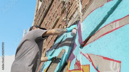 A man is painting a wall with graffiti. The wall is blue and the man is wearing a gray shirt