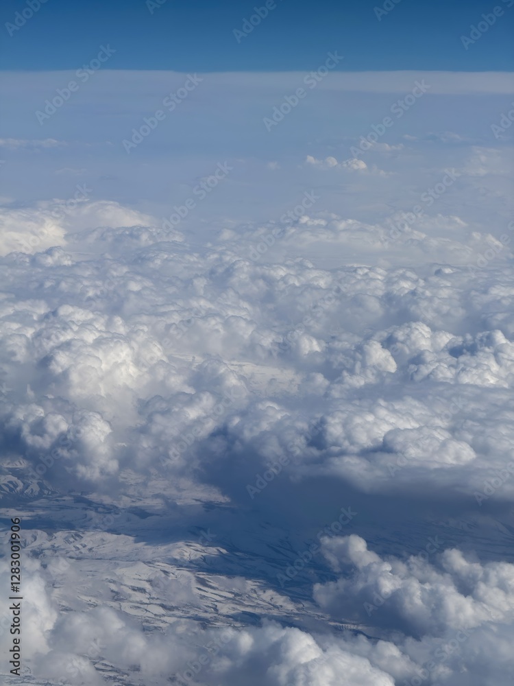 Aerial view of a snow-covered mountainous landscape partially obscured by thick, fluffy clouds. The sky above is a gradient of blue, contrasting with the white and shadowed areas of the clouds. Aerial view of a snow-covered mountainous landscape partially obscured by thick, fluffy clouds. The sky above is a gradient of blue, contrasting with the white and shadowed areas of the clouds.