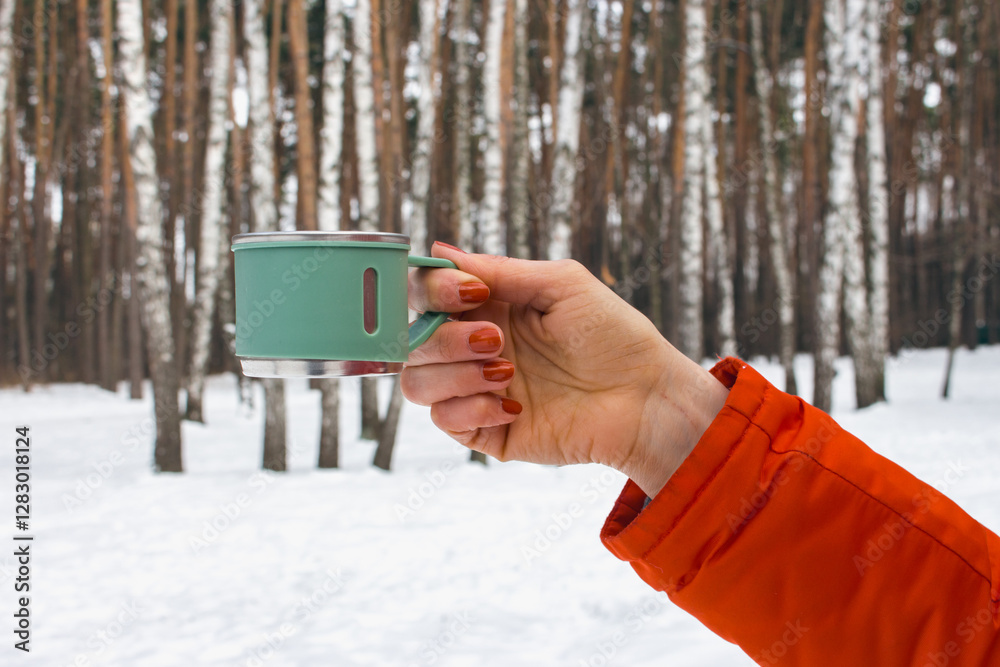 Woman in orange coat hanging mug in winter forest. Stainless cup for adventure trips in woman hand. Hand with thermo cup on winter landscape background. Winter tourism.