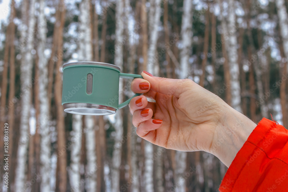 Woman in orange coat hanging mug in winter forest. Stainless cup for adventure trips in woman hand. Hand with thermo cup on winter landscape background. Winter tourism.