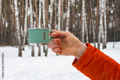 Obraz na płótnie Woman in orange coat hanging mug in winter forest. Stainless cup for adventure trips in woman hand. Hand with thermo cup on winter landscape background. Winter tourism. w salonie