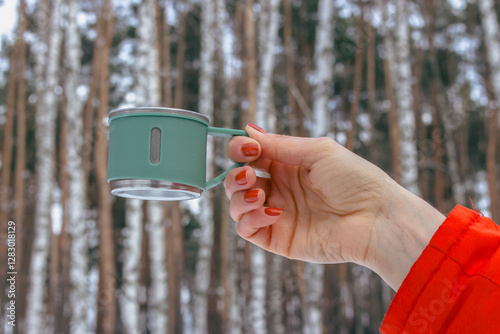 Obraz na płótnie Woman in orange coat hanging mug in winter forest. Stainless cup for adventure trips in woman hand. Hand with thermo cup on winter landscape background. Winter tourism. w salonie