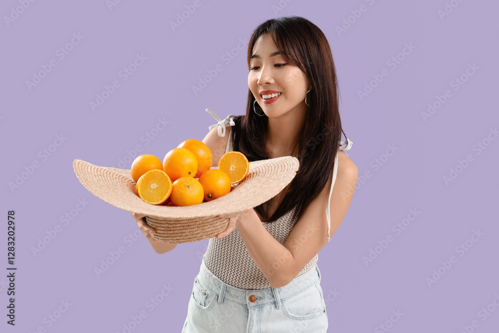 Young Asian woman with hat full of fresh oranges on lilac background