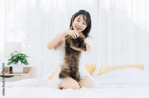 Asian happy young female sitting on white bed owner smiling holding playing fluffy tortoiseshell cute little kitten breed pussycat pet friend in bedroom at home, Concept love animal comfort rest care.