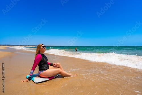 Beautiful mature woman sitting on bodyboard on ocean shore on sunny beach. Side view. Water sports in summertime photo