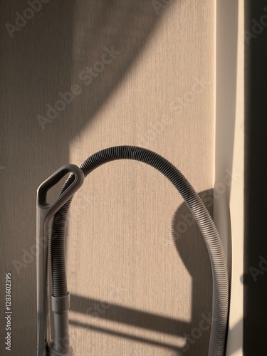 The household vacuum cleaner is near the wall.
Only the handle and hose of the device are visible. The wall is flooded with sunlight. Home cleaning equipment.