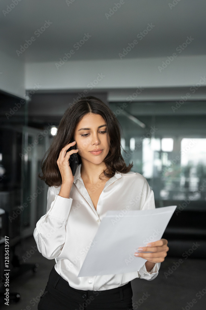 Fototapeta premium Middle aged latin businesswoman having business phone call with partners, clients, looking at documents. Young Hispanic woman financial analyst discussing tax report by cellphone in office. Vertical