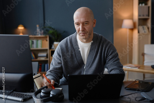 Wallpaper Mural Medium shot of mature man with shaved head writing records on online meeting in notebook, while using laptop at desk in cozy home office captured against blue wall Torontodigital.ca