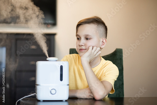 Cute teenager kid boy sitting near a humidifier with outgoing steam in a room.