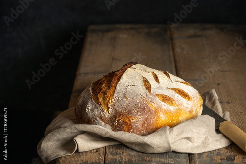 A homebaked artisan sourdough bread with scoring leaves on a teacloth next to a breadknife.