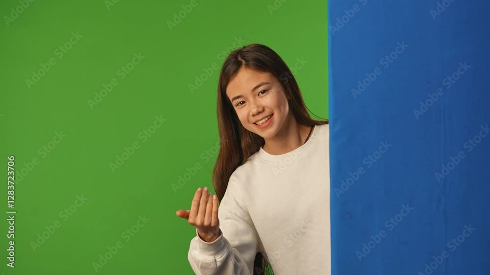 Asian girl child peeks out and waves hand, shows come here gesture, positive expression, isolated on chroma key green screen background mockup. Asian girl child peeks out and waves hand, shows come here gesture, positive expression, isolated on chroma key green screen background mockup.