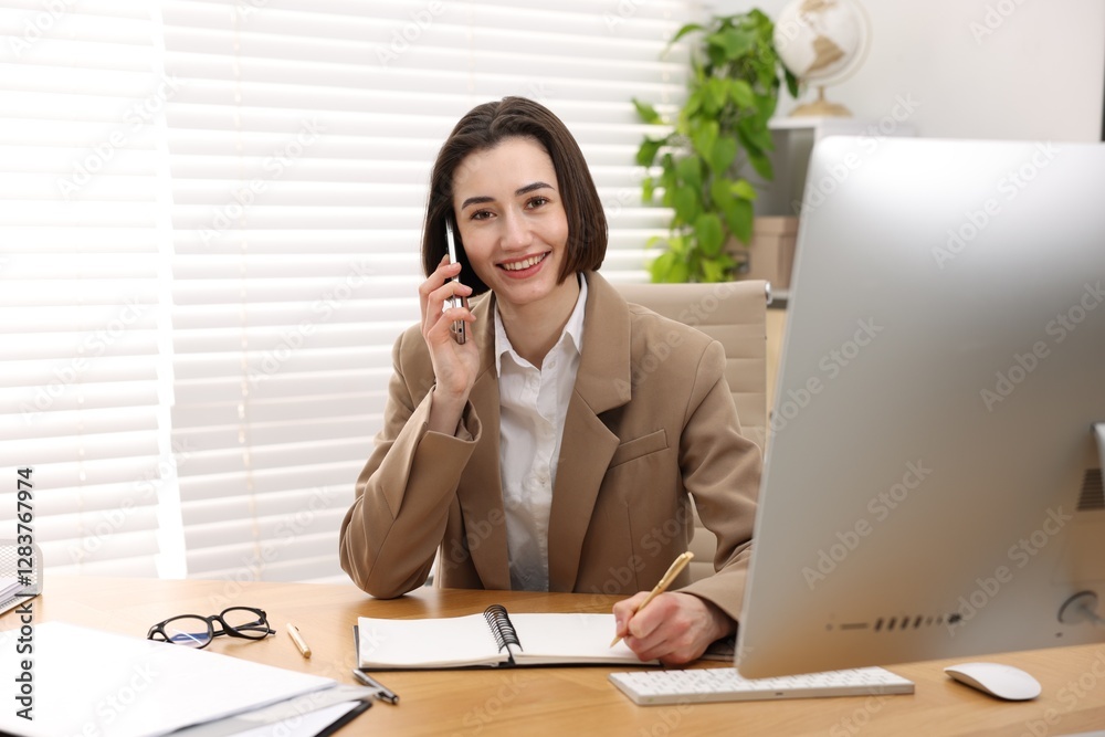 Smiling secretary talking on smartphone and taking notes at table in office