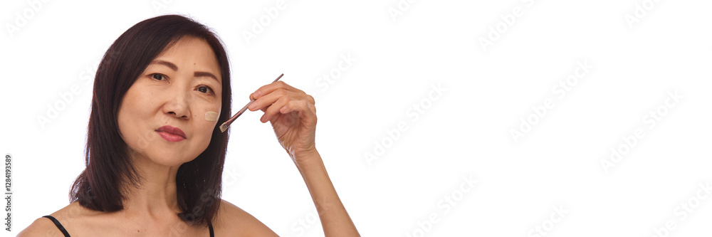 Portrait of mature Korean woman in her 60s with well-kept skin applying foundation or bb cream on her face with brush isolated on white background. Concept of skin care, makeup products, cosmetology Portrait of mature Korean woman in her 60s with well-kept skin applying foundation or bb cream on her face with brush isolated on white background. Concept of skin care, makeup products, cosmetology