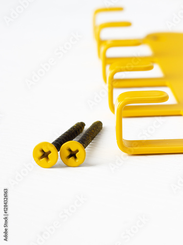 Close-up of two yellow screws with Phillips heads lying on a white surface next to a yellow metal wall-mounted hook rack. 