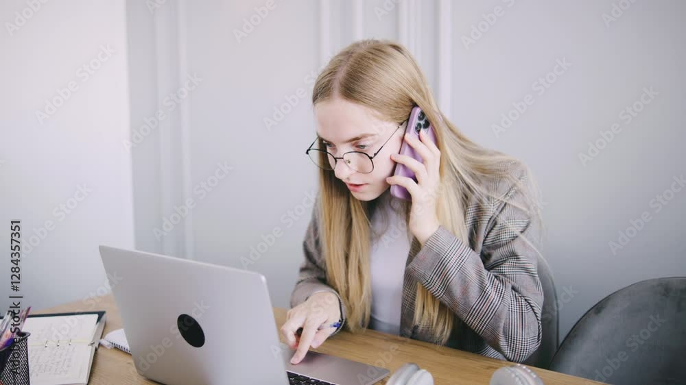 Young woman engaged in a phone conversation while working on a laptop at a modern workspace during the day Young woman engaged in a phone conversation while working on a laptop at a modern workspace during the day