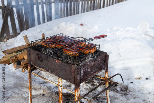 Winter, snow and sunny day. Rusty barbecue with firewood and hot dogs roasting on the grill. Delicious smoke.