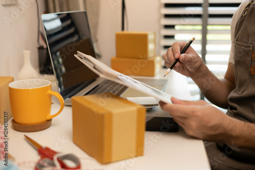 A man is sitting at a desk with a laptop and a clipboard. He is looking at a spreadsheet on the laptop and writing on the clipboard
