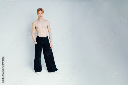 Handsome Male Model Stands Topless Against a White Background, Confidently Posing with Hands in the Pockets of Black Jeans, Looking at the Camera with a Serious and Neutral Facial Expression.