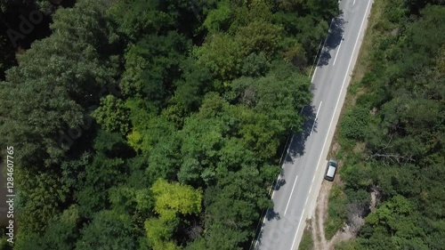 Aerial shot of a black car speeding down a road through the forest. On the left is a river, on the right is a parked gray car. Tracking shot