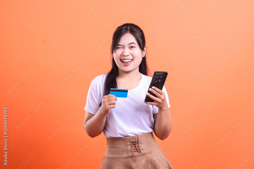 Excited young woman holding a credit card and a smartphone, winking, ready to shop online, isolated on orange background Excited young woman holding a credit card and a smartphone, winking, ready to shop online, isolated on orange background