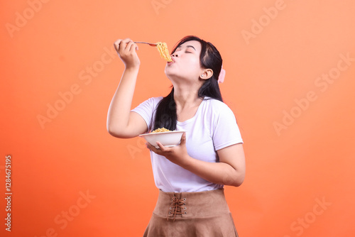 A young woman is happily eating noodles from a bowl with a fork, looking satisfied with closed eyes, isolated on orange background