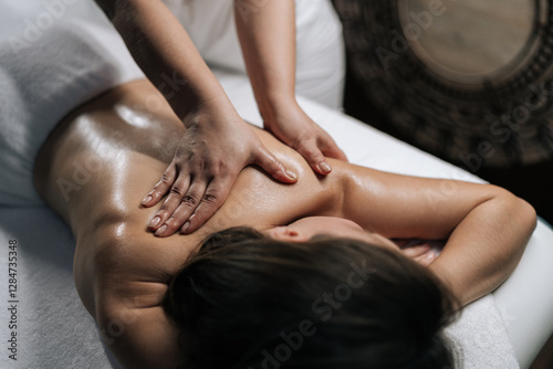 Closeup of massage therapist rubbing moisturizing oil into client shoulder and back during therapeutic treatment at serene wellness spa, targeting muscle tension and promoting relaxation