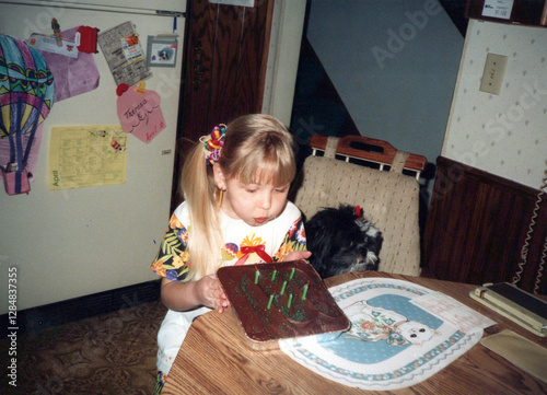Circa 1990 - Child (girl) blows out candles on her chocolate birthday cake with her pet dog. Vintage archival photo