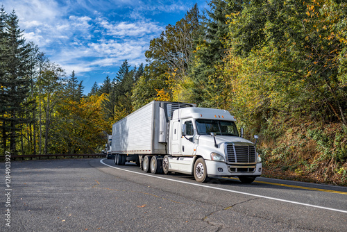 Low cab profile white big rig semi truck transporting cargo in refrigerator semi trailer running on the winding mountain road with autumn forest on the hillsides