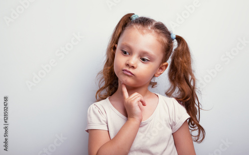 Happy fun cynical thinking kid girl with finger holding under the face looking down with empty copy space grey blue studio background. Closeup expression face portrait