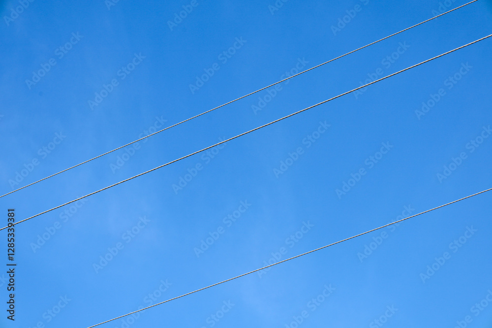 Two power lines are shown stretching horizontally across the frame against a bright blue sky. The lines are thin, dark in color, clearly distinguishable against the sky. Two power lines are shown stretching horizontally across the frame against a bright blue sky. The lines are thin, dark in color, clearly distinguishable against the sky.