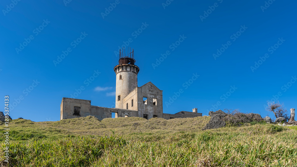An old lighthouse on a tropical island. A dilapidated building with a cylindrical tower, empty window openings against the blue sky. The green grass on the hill. Mauritius. Île aux Fouquets An old lighthouse on a tropical island. A dilapidated building with a cylindrical tower, empty window openings against the blue sky. The green grass on the hill. Mauritius. Île aux Fouquets