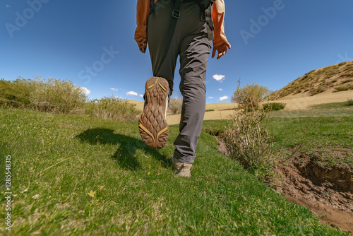 A man in bright clothes and a backpack on his back walks through the green grass against the background of the sands and defiantly shows the sole of his shoes. Hiking shoes and clothes on a background photo