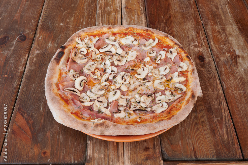 A pizza with mushrooms and ham on it sits on a wooden table A pizza with mushrooms and ham on it sits on a wooden table