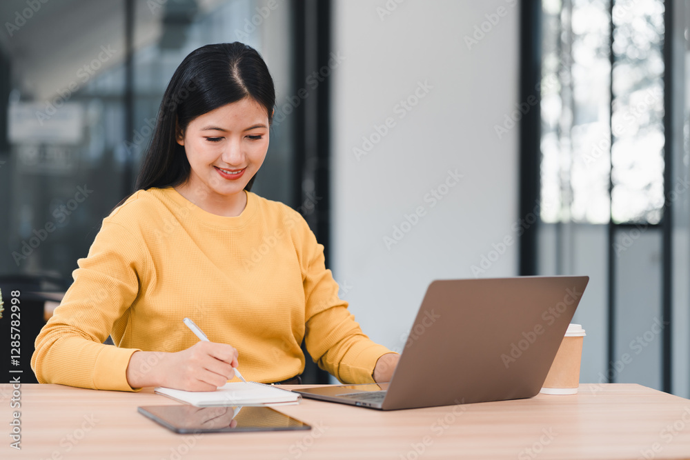 Woman in yellow sweater working on laptop while taking notes in notebook at modern office desk. Woman in yellow sweater working on laptop while taking notes in notebook at modern office desk.