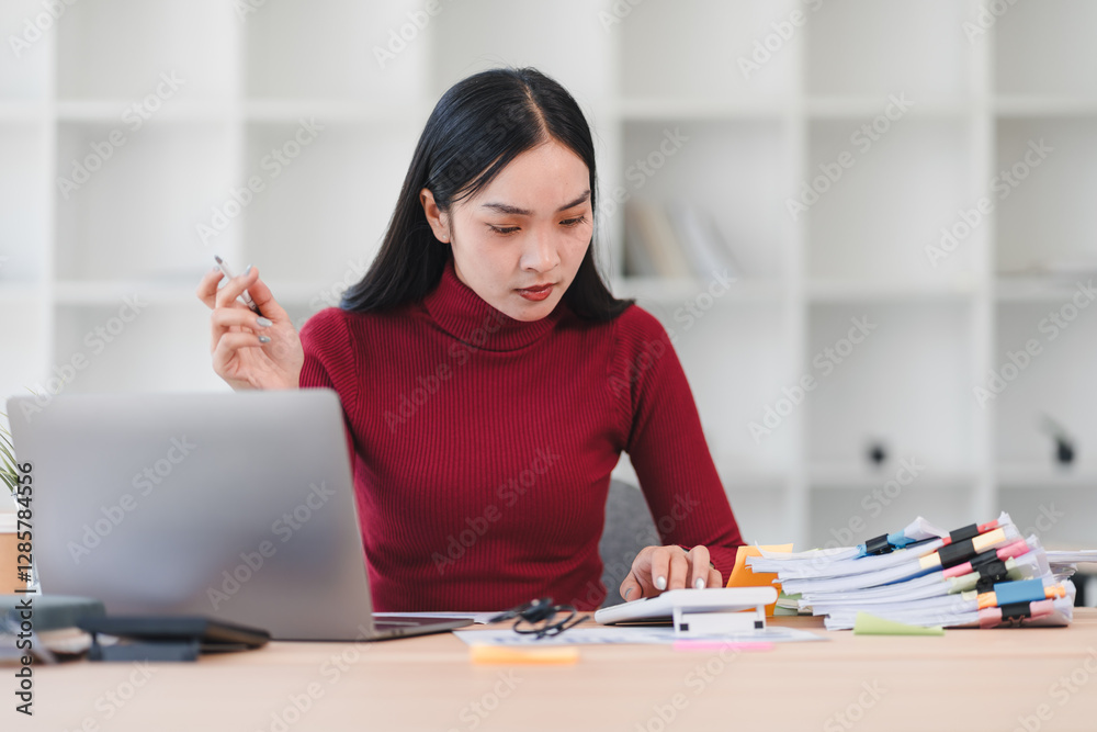 Focused Asian woman in red sweater working at desk with laptop, calculator, and stacks of documents. Focused Asian woman in red sweater working at desk with laptop, calculator, and stacks of documents.