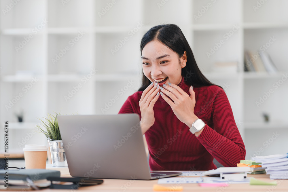Woman in red sweater expresses excitement while working on laptop at desk in bright office. Woman in red sweater expresses excitement while working on laptop at desk in bright office.